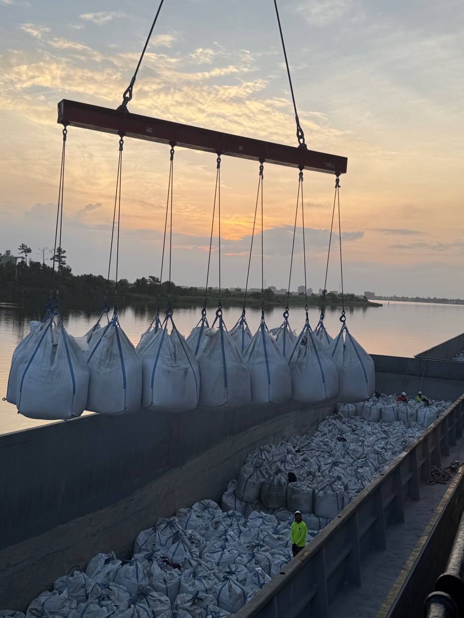 GCS Industrial Group cement supersack discharge — crane spreader bar lifting a row of bulk cement bags at sunset on the Gulf Coast waterway